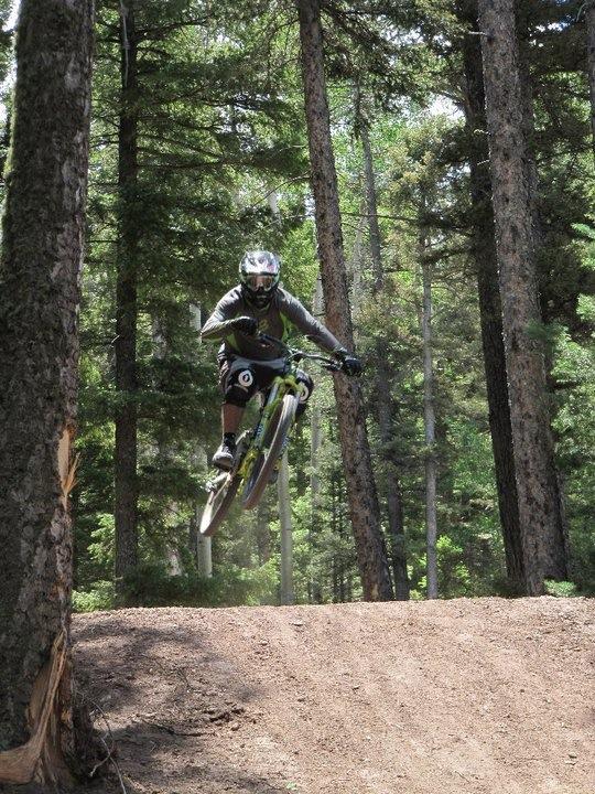 A mountain biker performing a jump on a dirt trail, surrounded by tall trees and greenery in a forest setting. The rider is wearing protective gear and is airborne above the trail. Angel Fire Bike Park mountain bike trail.