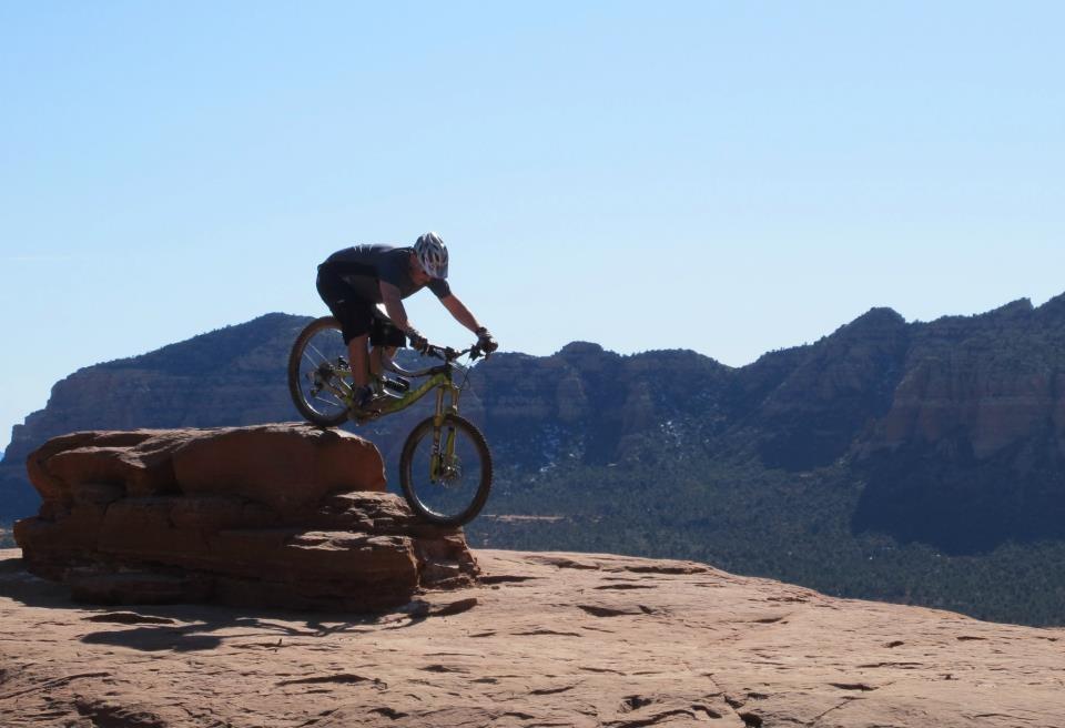 A mountain biker performing a stunt on a large rock formation, with a backdrop of mountains and clear blue sky. The biker is positioned mid-air, demonstrating balance and skill. Bell Rock Area Trails mountain bike trail.