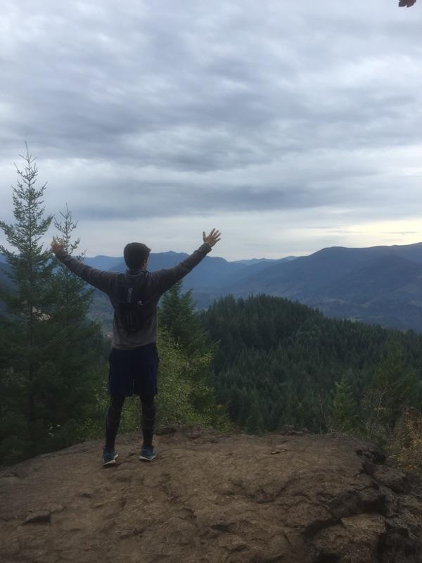 Santa Cruz Blur: A person standing on a rocky outcrop with arms raised, overlooking a vast mountainous landscape filled with evergreen trees under a cloudy sky.
