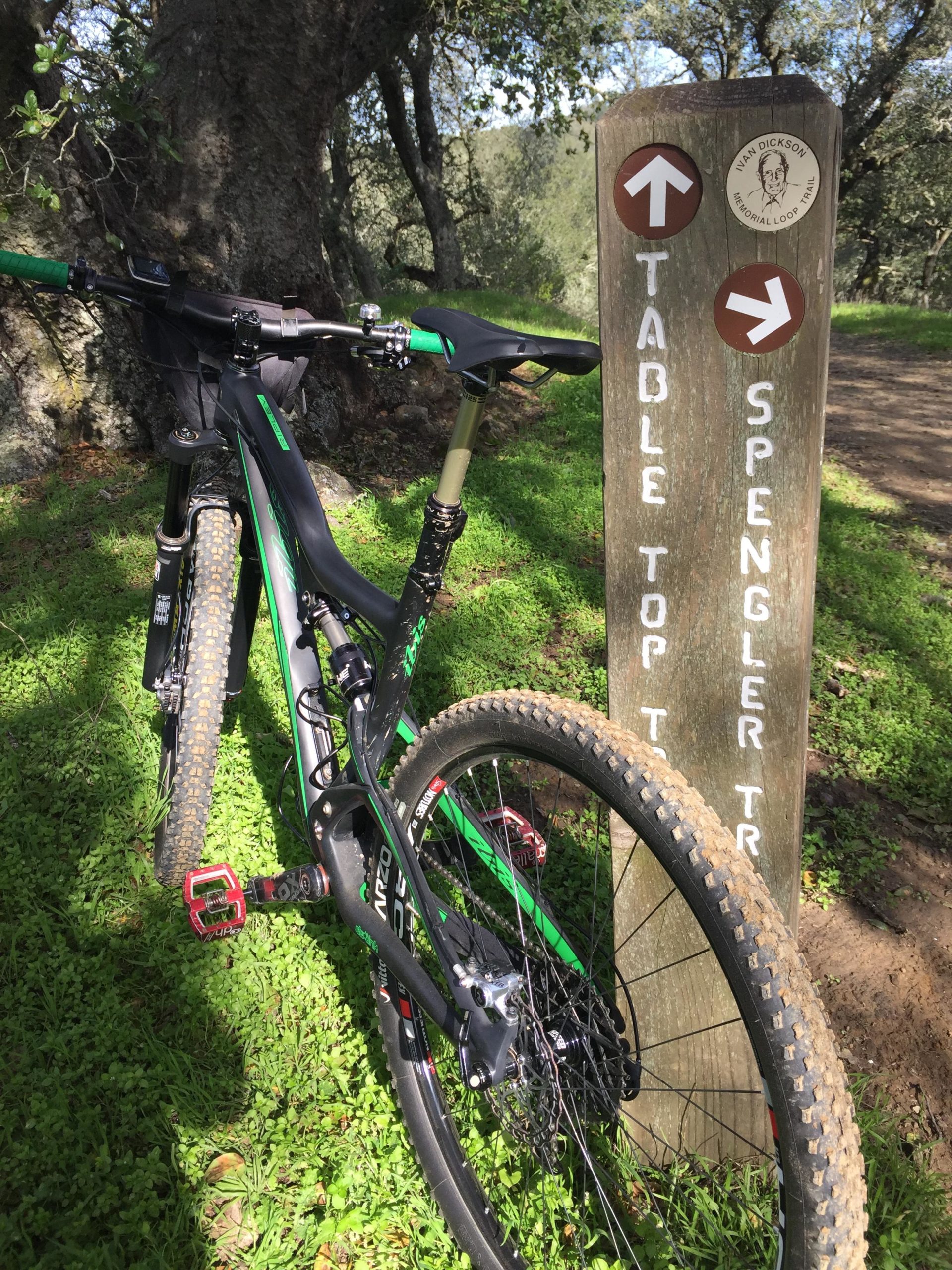 A mountain bike leaning against a wooden trail sign indicating directions for the Table Top Trail and Spengler Trail. The surrounding area features lush green grass and trees in a natural setting. Briones Regional Park mountain bike trail.