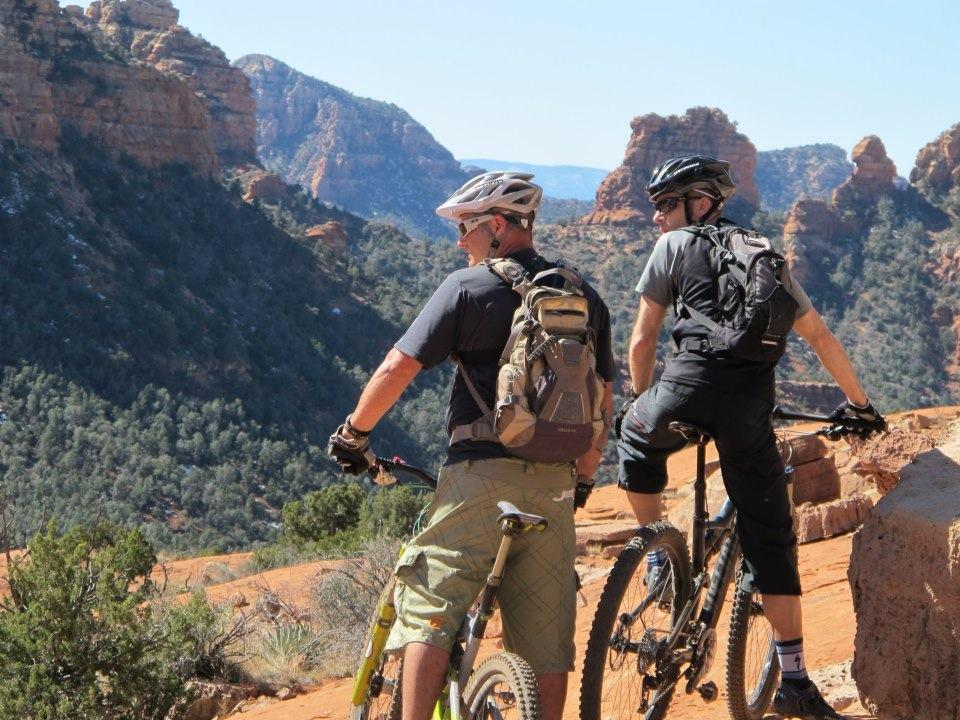 Alt tag: Two mountain bikers, one wearing a helmet and a backpack, stand beside their bikes while overlooking a mountainous landscape with red rock formations and greenery. Bell Rock Area Trails mountain bike trail.