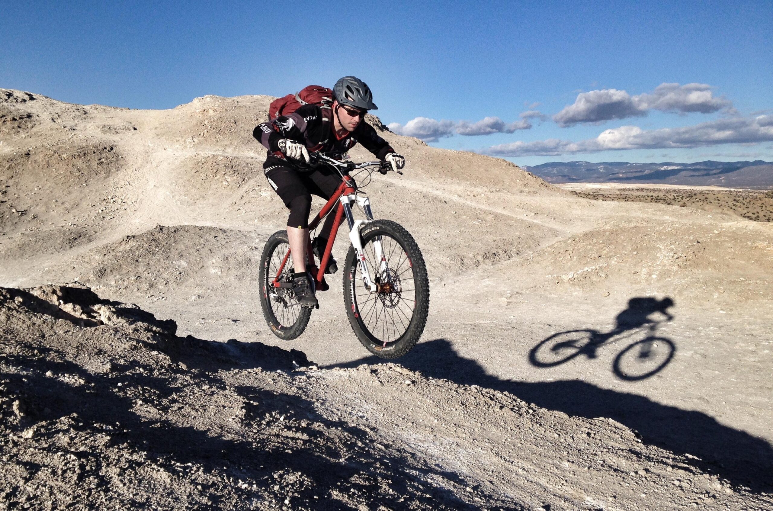 A mountain biker jumping over a rocky terrain, with a clear blue sky in the background. The biker is wearing a helmet and protective gear, riding a red bike, and casting a shadow on the ground below. The landscape around features dusty hills and sparse vegetation. White Ridge Bike Trails mountain bike trail.