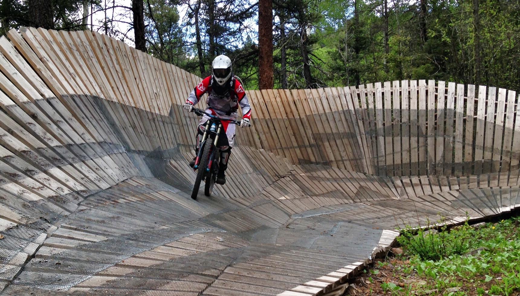 A mountain biker rides along a wooden pump track nestled in a forested area, showcasing skill and agility on the curved, textured surface. The track features layered wooden boards and steep banks, surrounded by trees and greenery. The biker is wearing a helmet and protective gear, emphasizing the action and excitement of the sport. Angel Fire Bike Park mountain bike trail.