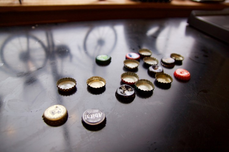 Close-up of various beer bottle caps scattered on a dark table surface, with a reflection of a bicycle in the background.