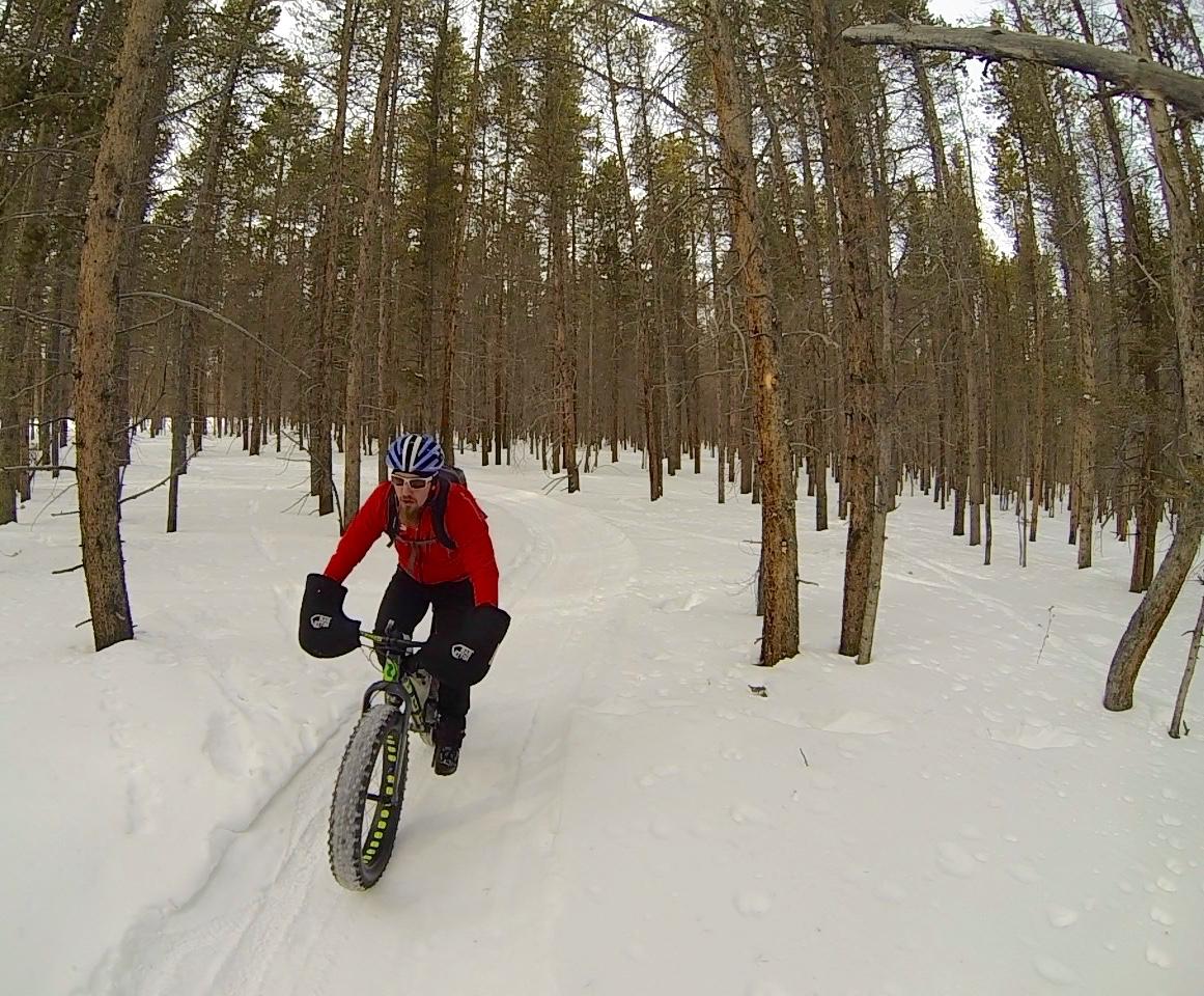 A person riding a fat bike through a snowy forest trail, surrounded by tall pine trees. The rider is wearing a red jacket and blue helmet, equipped with handlebar mitts for warmth. Colorado Mountain College Trails mountain bike trail.