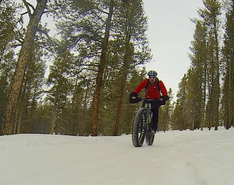 A cyclist riding a fat bike on a snow-covered trail in a forest, surrounded by tall pine trees. The rider is wearing a red jacket and a helmet, with snow gently falling in the background. Colorado Mountain College Trails mountain bike trail.