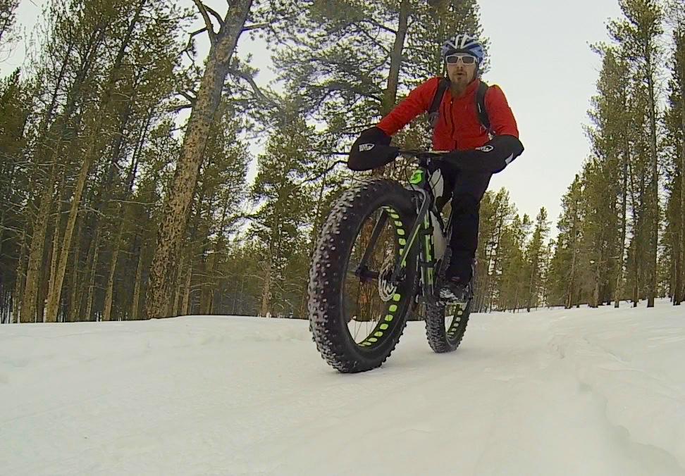 A person riding a fat bike on a snowy trail in a forest, wearing a red jacket and sunglasses. Pine trees are visible in the background, and the snow-covered ground shows tire tracks. Colorado Mountain College Trails mountain bike trail.