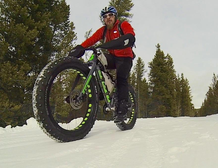 A cyclist riding a fat bike on a snow-covered trail, dressed in a red jacket, black pants, and a helmet, with trees in the background. Colorado Mountain College Trails mountain bike trail.