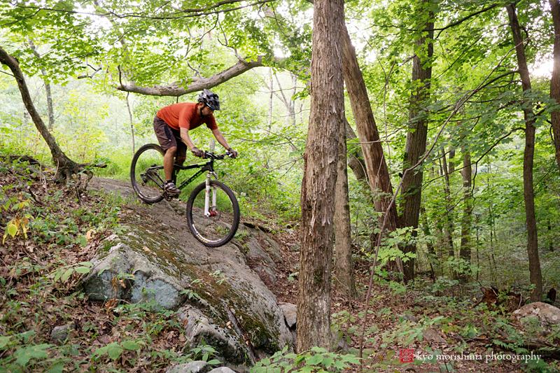 A mountain biker navigating a rocky trail in a dense forest, wearing a helmet and an orange shirt. Sunlight filters through the trees, illuminating the greenery around the rider as they maneuver over a large rock. Allamuchy State Park-North mountain bike trail.