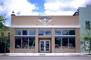 A storefront with large windows and a decorative sign featuring a winged design, set against a blue sky with clouds. The building has a beige facade with a modern aesthetic, flanked by two neighboring buildings.