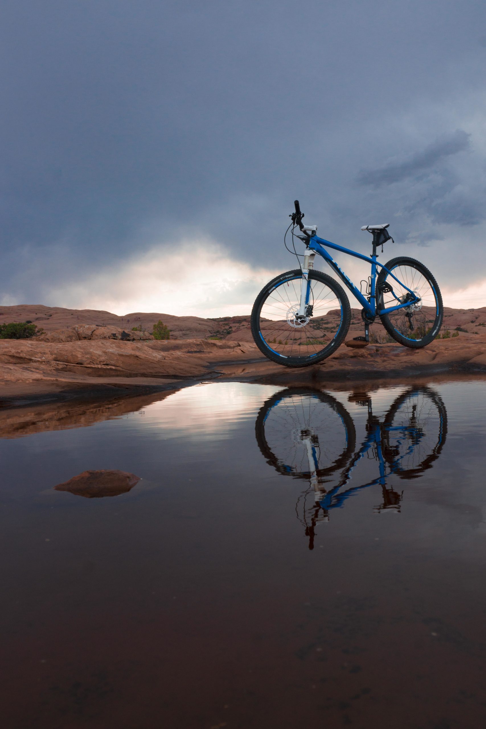 A blue mountain bike stands on rocky terrain, reflecting in a still puddle of water. Dark clouds loom overhead, creating a dramatic sky in the background. Slickrock mountain bike trail.