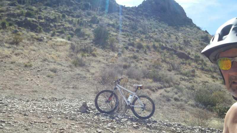 A cyclist wearing a helmet and sunglasses poses for a selfie, with a mountain bike parked on rocky terrain in the background. A dry, mountainous landscape with sparse vegetation is visible under a clear sky. Dona Ana mountain bike trail.