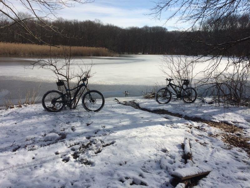 Two bicycles parked on a snowy shore beside a partially frozen lake, with trees and reeds in the background under a clear blue sky. Wolfes Pond park mountain bike trail.