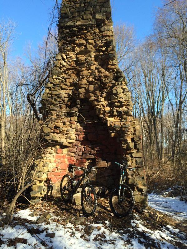Two mountain bikes are leaning against a tall stone ruin in a forested area. The structure, which appears weathered and partially collapsed, is made of large stones and has patches of red brick visible. In the background, trees without leaves are visible against a clear blue sky, with some patches of snow on the ground. Trails seperated by streets mountain bike trail.