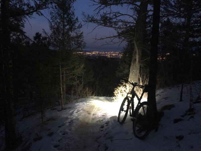 A mountain bike is leaning against a tree on a snowy trail, illuminated by a dim light as dusk settles. In the background, the glow of a city skyline is visible, contrasting with the darkening sky and surrounding trees. Cheyenne Mountain State Park mountain bike trail.