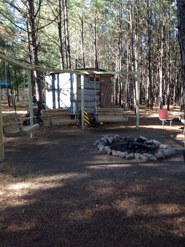 A peaceful outdoor setting in a forest, featuring a small wooden shelter with a sign reading "Bike Trail," wooden benches for seating, and a fire pit surrounded by stones. The area is surrounded by tall pine trees and dappled sunlight. Mt. Zion Bike Trails mountain bike trail.