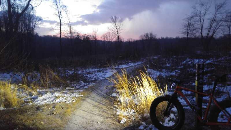 A dimly lit trail through a winter landscape, with patches of snow on the ground. A bright light illuminates the pathway and surrounding grasses, while a mountain bike with an orange frame rests to the right. In the background, trees stand silhouetted against a colorful sky at dusk, suggesting a serene outdoor scene. Wissahickon Valley Park mountain bike trail.