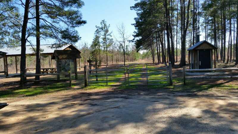 A clear view of a scenic outdoor area featuring a dirt path leading through a green space enclosed by wooden fencing. On the left, there are picnic shelters with benches, and on the right, a gated entrance to a wooded trail. Tall pine trees surround the area, and the sky is bright and clear, suggesting a sunny day. Lines Tract mountain bike trail.