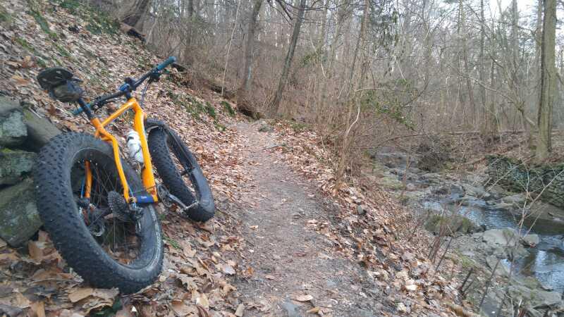 A bright orange fat bike rests against a stone wall beside a winding dirt path covered in fallen leaves. A small stream runs alongside the trail, surrounded by bare trees, indicating early spring or late autumn. Wissahickon Valley Park mountain bike trail.