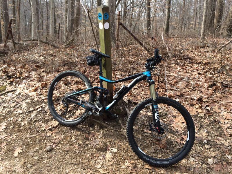 A mountain bike leaning against a wooden trail marker in a forest setting, surrounded by fallen leaves and trees. The trail marker displays blue and white circular symbols. White Clay Creek mountain bike trail.