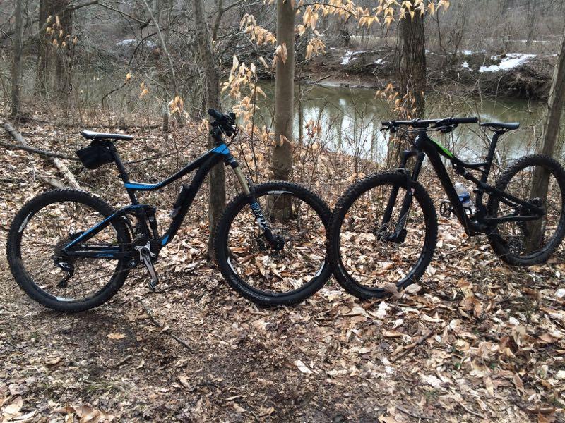 Two mountain bikes are parked side by side on a leaf-covered forest trail, with a creek visible in the background. The bike on the left features a black frame with blue accents, while the one on the right has a predominantly black design with green details. The surrounding trees are bare, indicating early spring, and some remnants of snow can be seen in the distance. White Clay Creek mountain bike trail.