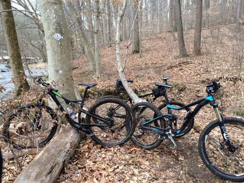 Two mountain bikes are parked next to a tree on a forest path covered with fallen leaves. A stream is visible in the background, and the trees are bare, indicating early spring or late fall. One bike is predominantly black with green accents, while the other features a combination of black and blue. White Clay Creek mountain bike trail.