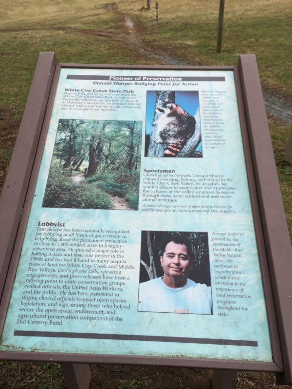 Image showing a historical information sign titled "Pioneer of Preservation" honoring Donald Sharpe. The sign includes sections about his contributions as a sportsman and lobbyist for environmental preservation, alongside images of a raccoon and a wooded trail. It highlights his work in promoting the protection of natural areas in White Clay Creek State Park and his efforts in conservation. White Clay Creek mountain bike trail.