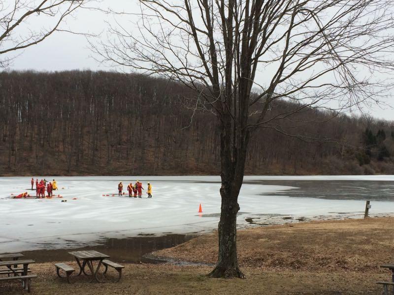 A group of rescue personnel in bright red and yellow uniforms are conducting a training exercise on a partially frozen lake, with a bare tree in the foreground and a wooded area in the background under overcast skies. Round Valley Recreation Area mountain bike trail.