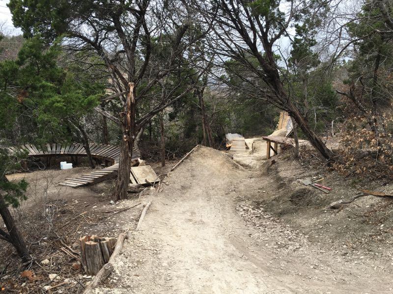 A dirt path winding through a wooded area, featuring wooden ramps and platforms on either side. Sparse trees and underbrush line the trail, which appears to be designed for biking or outdoor activities. The scene is overcast, creating a muted atmosphere. Big Cedar Wilderness Trails mountain bike trail.