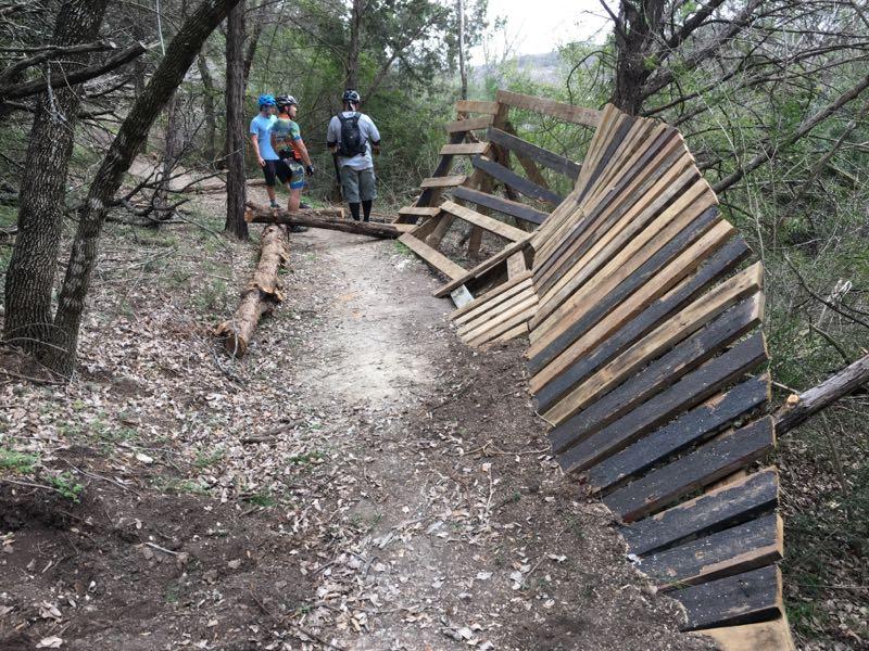Three mountain bikers stand on a dirt trail surrounded by trees, observing a wooden ramp structure that is leaning and partially collapsed. A log is positioned across the trail, adding to the natural setting. The scene is set in a wooded area, with fallen leaves and vegetation along the path. Big Cedar Wilderness Trails mountain bike trail.