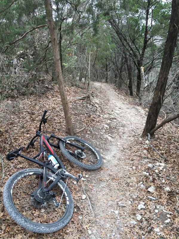 A mountain bike lying on its side on a dirt trail surrounded by trees and fallen leaves, with a winding path visible in the background. Big Cedar Wilderness Trails mountain bike trail.