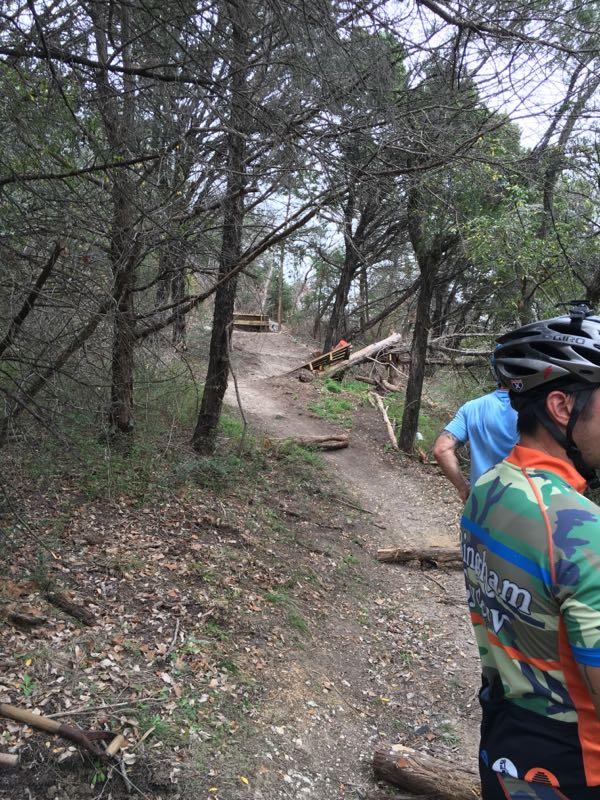 A winding dirt trail through a wooded area, with cyclists preparing to ride. One cyclist is wearing a brightly colored jersey and a helmet, while another is visible in the background. The path includes wooden obstacles and is surrounded by trees and scattered leaves. Big Cedar Wilderness Trails mountain bike trail.