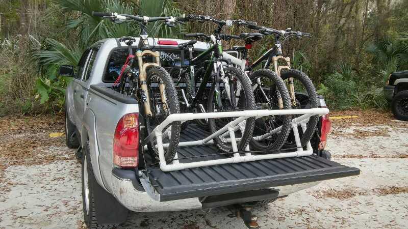A silver pickup truck parked in a forested area with a bike rack in the bed, holding four mountain bikes. The bikes are secured upright, with their wheels aligned and handlebars visible, featuring various colors. Lush greenery is seen in the background, providing a natural setting. Chuck Lennon Park mountain bike trail.
