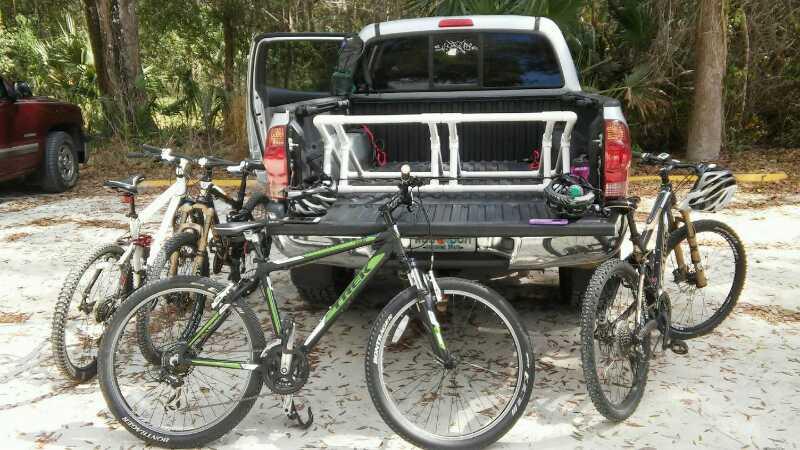 A black pickup truck parked on a sandy area with several mountain bikes secured in the truck bed and around the vehicle. The scene is set in a wooded area, with trees in the background. The bikes vary in color and style, showcasing their different features. Chuck Lennon Park mountain bike trail.