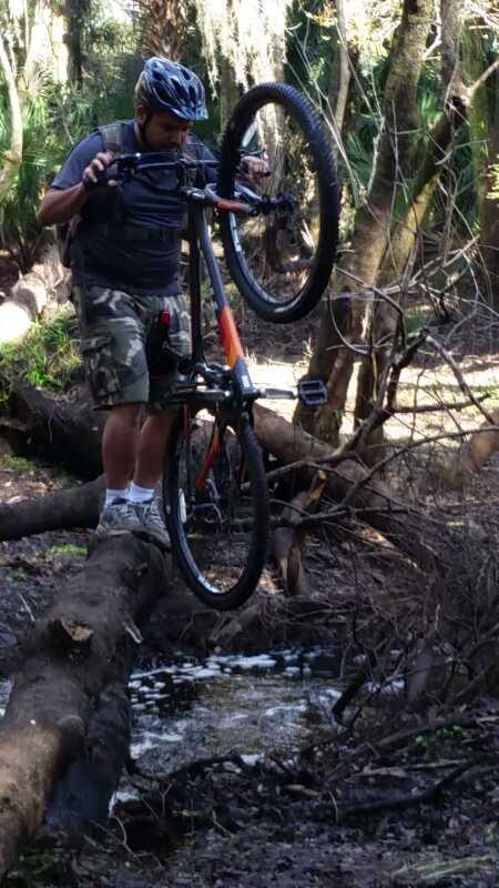 A cyclist performing a wheelie on a mountain bike while balancing on a fallen log over a stream in a wooded area. The rider is wearing a helmet and casual clothing, surrounded by lush greenery and a natural landscape. Little Big Econ State Forest mountain bike trail.