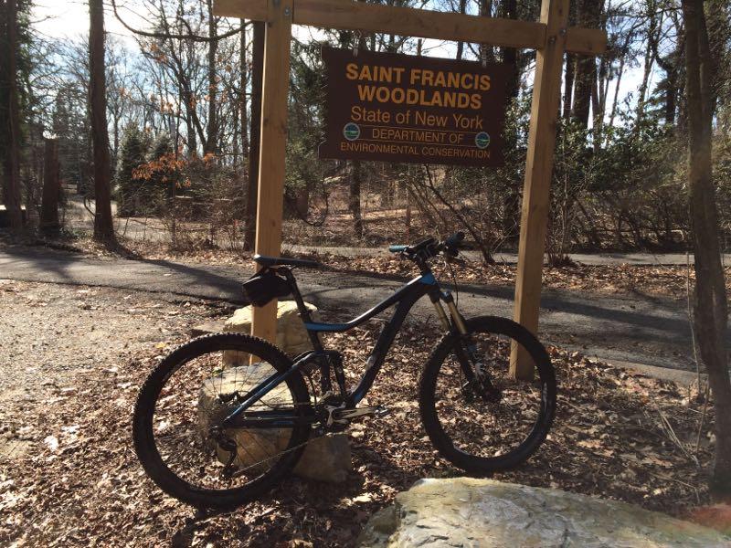 A mountain bike is parked next to a wooden sign that reads "Saint Francis Woodlands, State of New York, Department of Environmental Conservation." The scene is set in a wooded area with fallen leaves on the ground and a pathway visible in the background. Richmond Avenue and Forest Hill road mountain bike trail.
