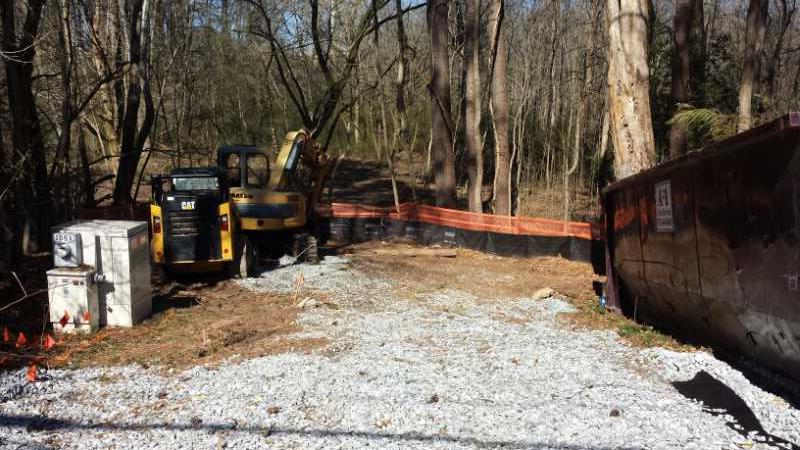 Construction site in a wooded area, featuring a yellow excavator and a concrete utility box. The ground is covered with gravel and bordered by orange safety fencing, with trees in the background. Ira B Melton mountain bike trail.
