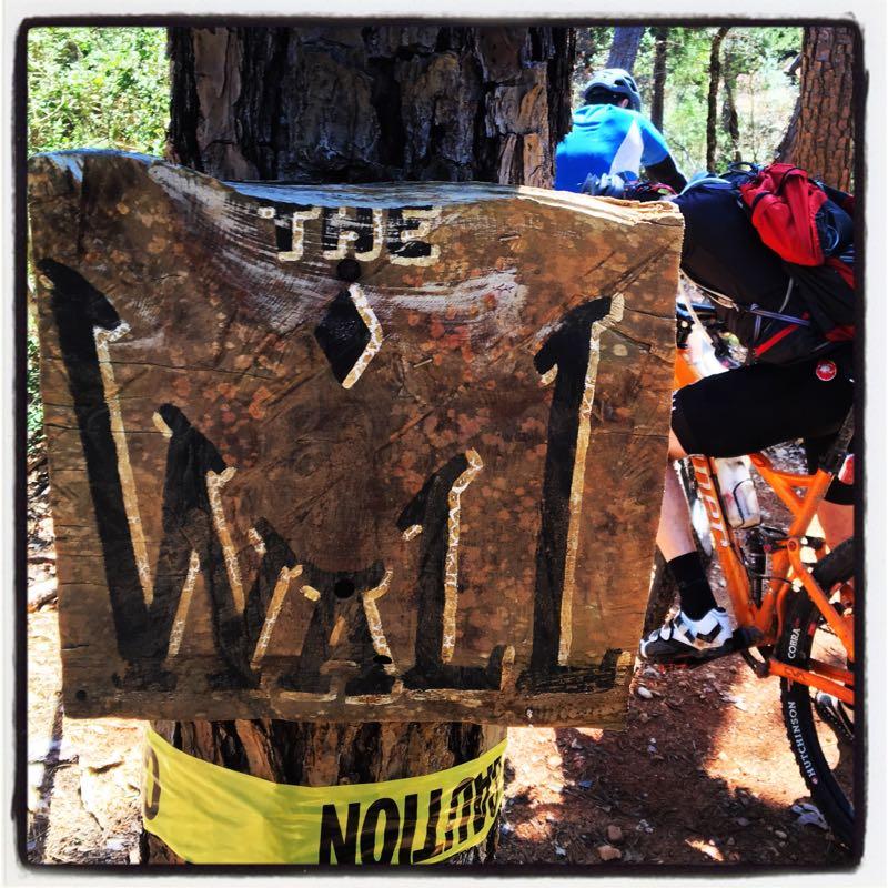 A weathered wooden sign reading "THE WALL" with arrows pointing upward, mounted on a tree, surrounded by a yellow caution tape. In the background, a person on an orange mountain bike is partially visible, riding along a dirt trail in a forested area. Rocky Hill Ranch mountain bike trail.
