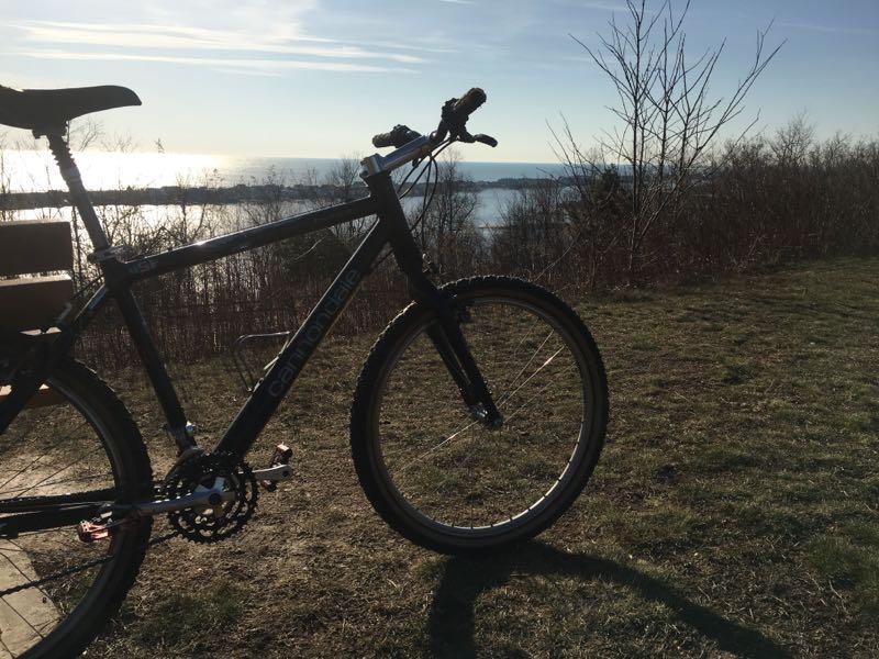 A black mountain bike stands on a grassy hillside overlooking a body of water. The sun is shining in a clear blue sky, reflecting off the water's surface. Sparse trees and shrubs are visible in the background, indicating a scenic outdoor landscape. Hartshorne Woods Park mountain bike trail.