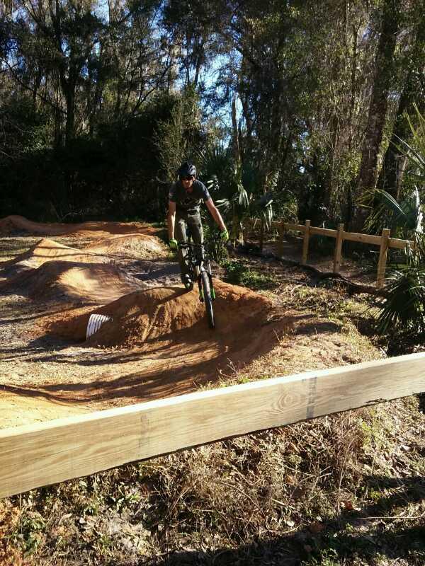 A person riding a mountain bike is mid-jump over a dirt ramp in a wooded area. The scene includes dirt mounds and a wooden barrier in the foreground, with lush greenery in the background. The rider is wearing a helmet and gloves, showcasing an active outdoor sport environment. Chuck Lennon Park mountain bike trail.