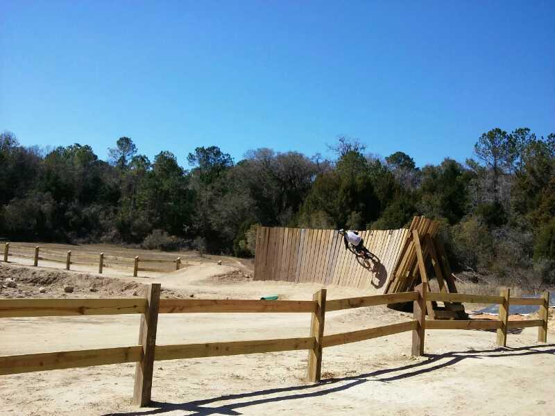 A cyclist performing a trick on a wooden wall ride at a dirt biking park, surrounded by trees and a clear blue sky. The scene includes a fenced area and a dirt pathway, showcasing a sunny day ideal for outdoor activities. Santos mountain bike trail.