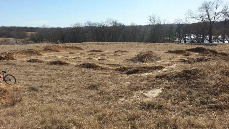A wide-open grassy area featuring several small earth mounds, with a bicycle partially visible on the left side of the image. The background shows sparse trees and a clear blue sky, indicating a rural landscape. Shawnee Mission Park mountain bike trail.
