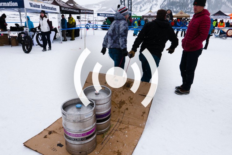 A snowy outdoor event scene featuring participants and tents, with two individuals pulling a cardboard ramp set up with two beer kegs on it. Spectators in the background are under canopies marked with "Visitor Information" and event signage, while snowflakes fall gently.