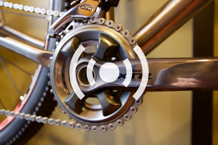 Close-up of a bicycle crankset featuring a Shimano XTR component. The image highlights the gears, chain, and polished metallic finish of the crank, set against a blurred background to emphasize the bike's intricate parts.