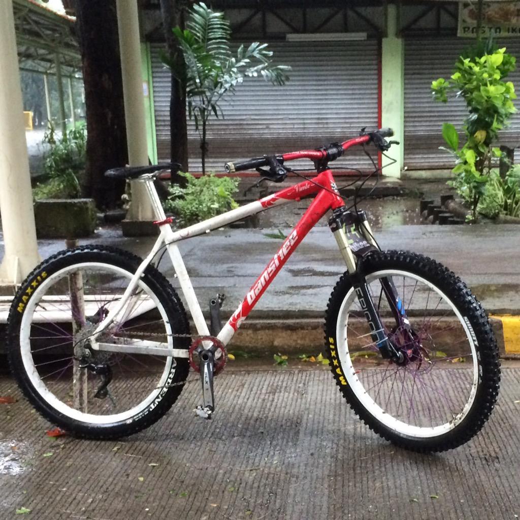 Banshee Viento: A mountain bike with a red and white frame, featuring thick, knobby tires, is parked on a textured pavement. In the background, there are blurred green plants and partially closed shutters of a building, suggesting an outdoor setting after rain.