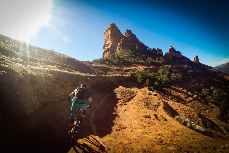 A mountain biker riding down a rocky, sunlit trail with reddish-brown terrain and towering rock formations in the background under a clear blue sky.