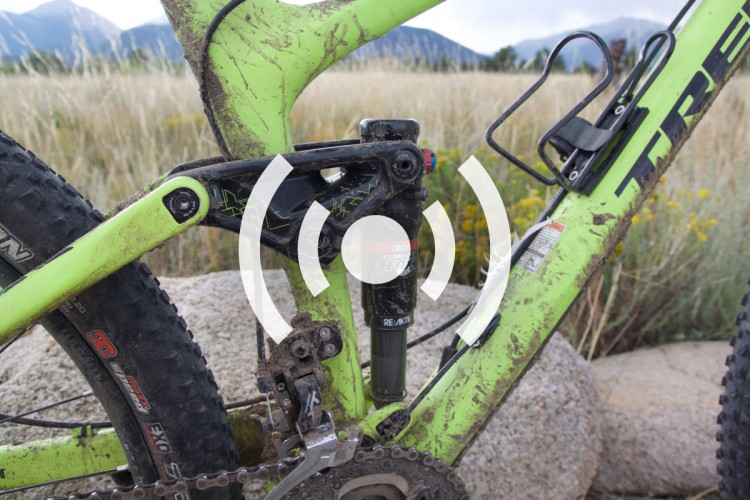 A close-up view of a green mountain bike frame with visible dirt and mud, showcasing the rear suspension and mechanical components. The background features a natural setting of tall grass and mountains.