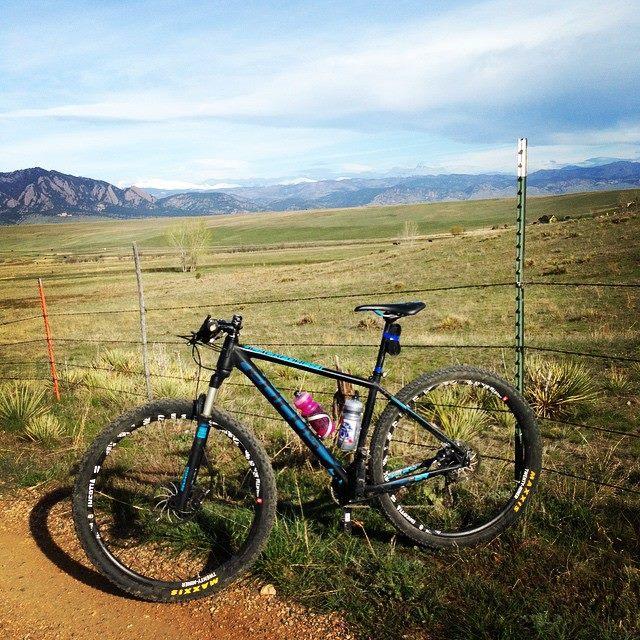 A mountain bike parked beside a fence on a scenic trail, with expansive green fields and mountains in the background under a clear blue sky. The bike has a water bottle attached. Marshall Mesa Area mountain bike trail.