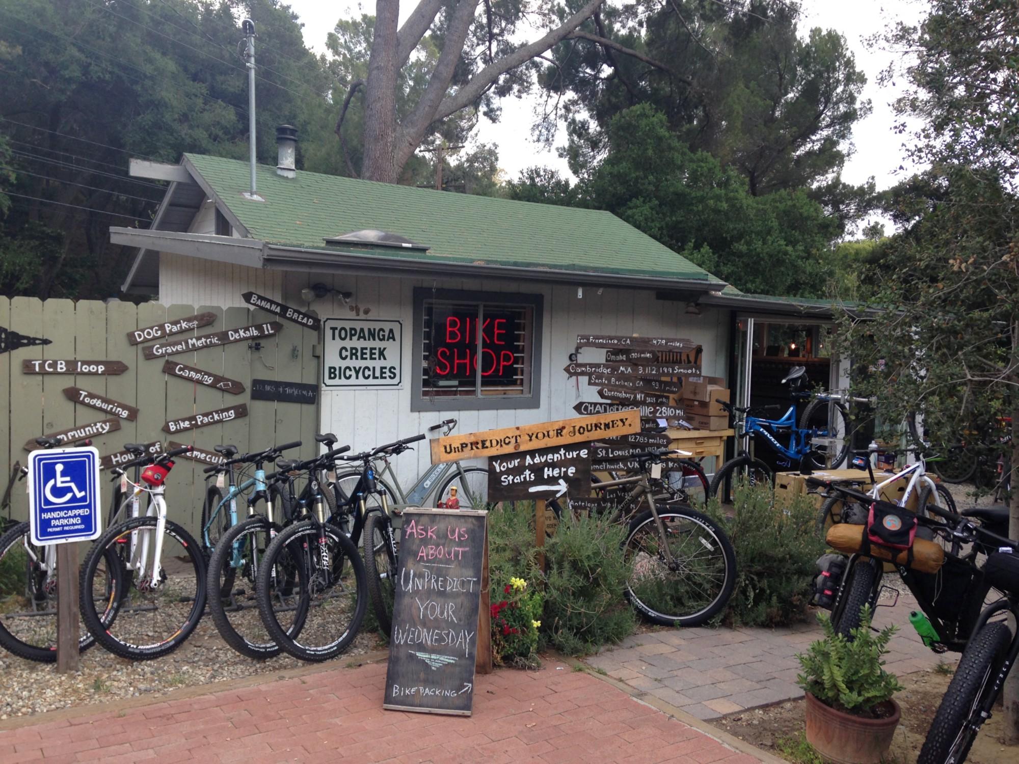 A quaint bike shop with a green roof, displaying various bicycles in front of a wooden fence adorned with directional signs for biking trails and destinations. A chalkboard sign promotes events and services, and there are plants around the entrance, creating an inviting atmosphere for cyclists.