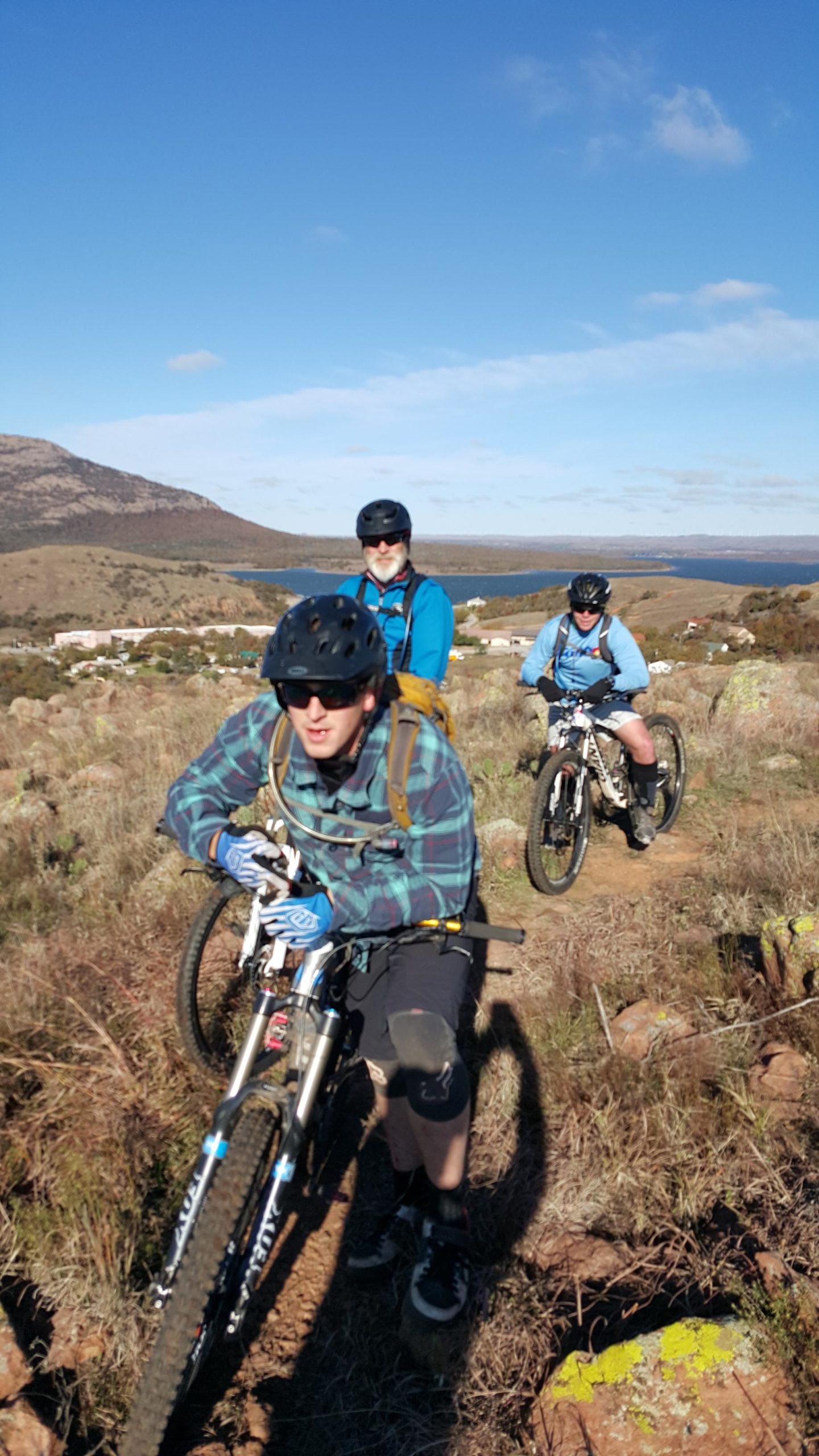 Three mountain bikers navigating a rocky trail surrounded by grass and shrubs, with a scenic landscape in the background that features hills and a lake under a blue sky. Lake Lawtonka Trails mountain bike trail.